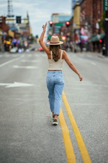 A woman wearing a hat and casual attire walks confidently down the middle of a city street with her hand raised in a peace gesture. The street is mostly empty, surrounded by blurred buildings and bright lights in the background.