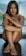 A person with long hair sits cross-legged, arms wrapped around their knees, with tattoos visible on both arms. The background is blurred, suggesting an indoor setting. The expression is calm and introspective.