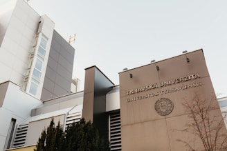 The image features a modern architectural building with a facade displaying the words 'Trnavská Univerzita Universitas Tyrnaviensis'. The structure is sleek with clean lines, large glass windows, and a light gray color palette. A leafless tree and some shrubbery are visible in the foreground, adding a natural element to the urban setting.