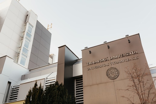 The image features a modern architectural building with a facade displaying the words 'Trnavská Univerzita Universitas Tyrnaviensis'. The structure is sleek with clean lines, large glass windows, and a light gray color palette. A leafless tree and some shrubbery are visible in the foreground, adding a natural element to the urban setting.