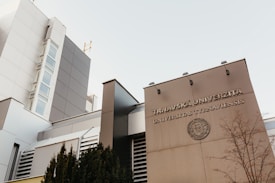 The image features a modern architectural building with a facade displaying the words 'Trnavská Univerzita Universitas Tyrnaviensis'. The structure is sleek with clean lines, large glass windows, and a light gray color palette. A leafless tree and some shrubbery are visible in the foreground, adding a natural element to the urban setting.