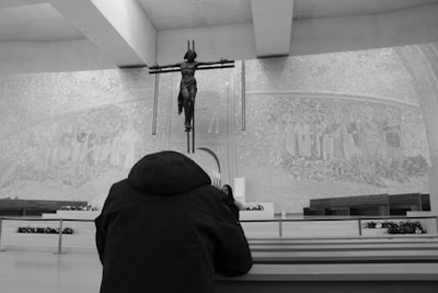 A person is kneeling in prayer before a large crucifix inside a modern chapel. The walls are adorned with detailed religious mosaics depicting saints or biblical figures. The space is minimalistic, focusing attention on the religious imagery and the act of devotion.