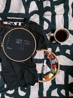 A black fabric with an embroidery hoop around a face pattern is placed on a printed black and white surface. Next to it, there is another embroidery hoop with colored threads wrapped around small cards and a black marker. A cup filled with dark liquid is placed to the right, likely coffee or tea.