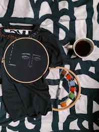 A black fabric with an embroidery hoop around a face pattern is placed on a printed black and white surface. Next to it, there is another embroidery hoop with colored threads wrapped around small cards and a black marker. A cup filled with dark liquid is placed to the right, likely coffee or tea.