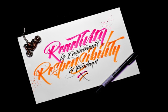 A piece of paper with colorful calligraphy reading 'Creativity is Contagious, Pass it on' written in vibrant pink and orange ink. The paper rests on a dark surface, with a black pen placed beside it. A motorcycle keychain sits on the top left corner of the paper.