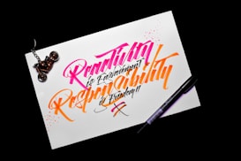 A piece of paper with colorful calligraphy reading 'Creativity is Contagious, Pass it on' written in vibrant pink and orange ink. The paper rests on a dark surface, with a black pen placed beside it. A motorcycle keychain sits on the top left corner of the paper.