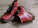 Two red leather boots with black laces are placed on a worn wooden floor. The boots show signs of wear and have a rugged appearance. In the background, there is a clear plastic bottle leaning against a textured wall.