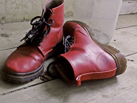Two red leather boots with black laces are placed on a worn wooden floor. The boots show signs of wear and have a rugged appearance. In the background, there is a clear plastic bottle leaning against a textured wall.