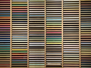 A collection of multi-colored sheets of paper organized in horizontal layers within a wooden shelving unit. The papers vary in hues including reds, blues, greens, yellows, and pastels, creating a visually striking array of colors.
