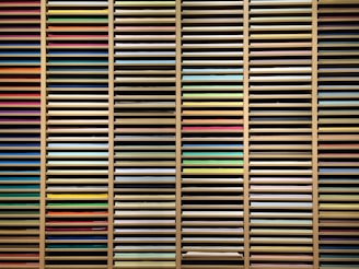 A collection of multi-colored sheets of paper organized in horizontal layers within a wooden shelving unit. The papers vary in hues including reds, blues, greens, yellows, and pastels, creating a visually striking array of colors.