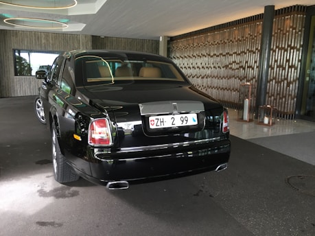 A luxurious black Rolls-Royce car is parked under a modern canopy with stylish circular lights above. The vehicle is positioned on a stone driveway, with wooden-panelled and glass walls in the background. The car's polished surface reflects its surroundings and the ambient lighting.