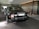 A luxurious black Rolls-Royce car is parked under a modern canopy with stylish circular lights above. The vehicle is positioned on a stone driveway, with wooden-panelled and glass walls in the background. The car's polished surface reflects its surroundings and the ambient lighting.