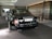 A luxurious black Rolls-Royce car is parked under a modern canopy with stylish circular lights above. The vehicle is positioned on a stone driveway, with wooden-panelled and glass walls in the background. The car's polished surface reflects its surroundings and the ambient lighting.