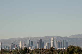 A city skyline featuring a range of modern skyscrapers is set against a backdrop of rocky mountains under a clear blue sky. The foreground includes lush green tree tops, adding a natural contrast to the urban landscape.