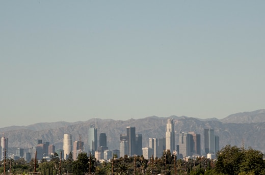 A city skyline featuring a range of modern skyscrapers is set against a backdrop of rocky mountains under a clear blue sky. The foreground includes lush green tree tops, adding a natural contrast to the urban landscape.