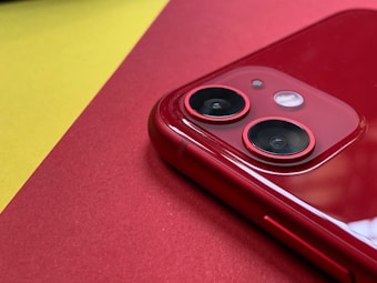 A close-up view of a red smartphone focusing on the dual camera lenses. The phone is placed on a red surface, with a contrasting yellow background. The shiny surface of the phone reflects light, emphasizing its glossy finish.