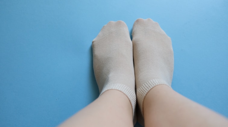 A pair of feet wearing light-colored socks are resting on a smooth, blue surface. The socks cover the feet entirely, showing a subtle texture and neutral tone that contrasts with the bright background. The feet are positioned close together with relaxed posture.