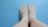 A pair of feet wearing light-colored socks are resting on a smooth, blue surface. The socks cover the feet entirely, showing a subtle texture and neutral tone that contrasts with the bright background. The feet are positioned close together with relaxed posture.