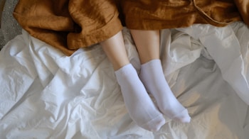 A pair of feet clad in white socks is seen resting on a crumpled white sheet. A brown blanket partially covers the legs, creating a cozy and relaxed setting.