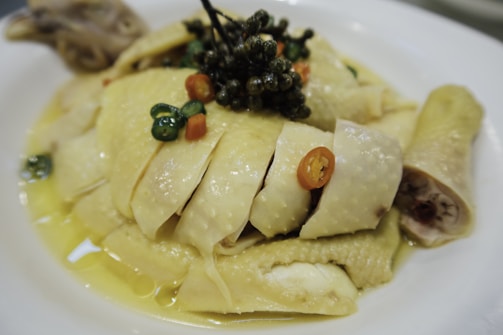 Sliced poached chicken served on a white plate, garnished with green peppercorns and small slices of red and green chilies. The chicken is bathed in a light yellow sauce, possibly oil or broth, giving it a glossy appearance.