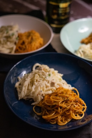 A close-up of three different plates of pasta, two featuring creamy white sauce and one with red sauce. The pasta is garnished with herbs and presented on dark blue and light-colored plates, with a bottle of wine visible in the background.