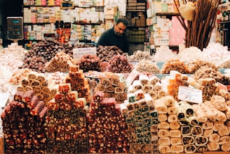 A market stall displaying a colorful and abundant assortment of sweets, including various types of Turkish delight arranged in stacks and piles. The sweets are diverse in color and texture, creating an eye-catching display. A vendor is seen in the background organizing the stall, surrounded by shelves filled with boxes and packages.