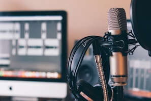A professional microphone with a pop filter is prominently featured in the foreground. Behind the microphone, a pair of headphones rests against a computer screen displaying audio waveforms, suggesting a recording or podcasting setup.