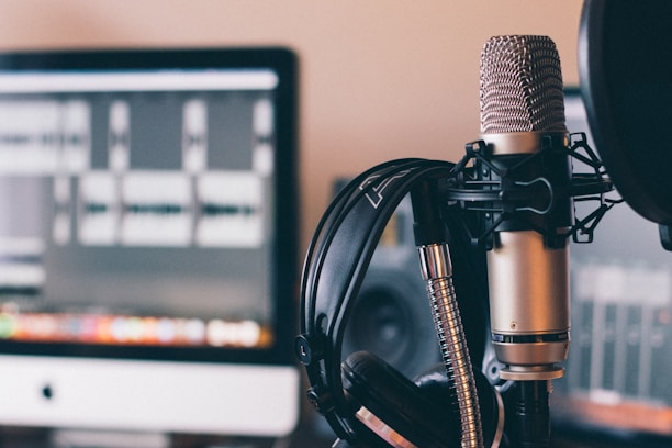 A professional microphone with a pop filter is prominently featured in the foreground. Behind the microphone, a pair of headphones rests against a computer screen displaying audio waveforms, suggesting a recording or podcasting setup.