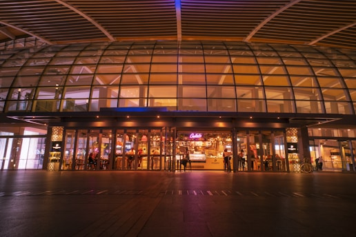 A modern architectural structure with a glass facade, featuring a warmly lit interior. People are seen sitting and standing inside what appears to be a café or bar, indicated by the neon signs and seating arrangements. The exterior has an expansive curved design with a combination of metal and glass panels.