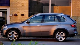 A sleek, modern SUV is parked on a street in an urban setting. The vehicle has a metallic silver exterior, prominent alloy wheels, and tinted windows. The backdrop consists of a yellow brick wall and glass windows of a building, reflecting a warm evening light.