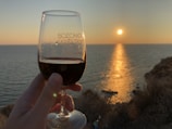 A hand holding a glass of red wine is positioned against the backdrop of a serene ocean view during sunset. The golden light of the setting sun casts a reflective path across the water, creating a peaceful and warm atmosphere. The glass has text printed on it.