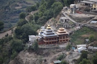 An ornate building with golden rooftops situated on a mountainside among green trees and surrounding smaller structures. The architecture is intricate with multiple tiers and appears to be a temple or religious site. The landscape is rugged with paths and terraces that wind through the hilly terrain.