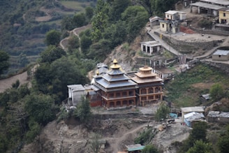 An ornate building with golden rooftops situated on a mountainside among green trees and surrounding smaller structures. The architecture is intricate with multiple tiers and appears to be a temple or religious site. The landscape is rugged with paths and terraces that wind through the hilly terrain.