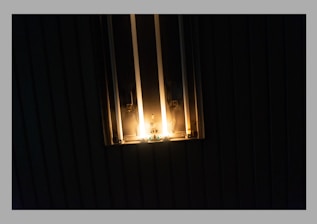 A dimly lit space features a wall-mounted fixture emitting a warm glow. The light casts subtle shadows, creating a cozy and intimate atmosphere. The setting is likely surrounded by darkness, emphasizing the brightness of the light source.