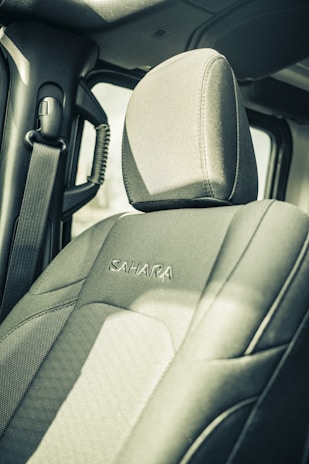 The interior of a vehicle featuring a front passenger seat with the word 'Sahara' embroidered on the fabric. The seatbelt hangs on the side, and part of the doorframe and window is visible. The image is captured in a muted, somewhat vintage style.