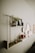 A minimalist wall shelf decorated with various items including a planter with green foliage, a framed photo, a glass terrarium, and small decorative objects like a pyramid. The shelf has a light wood finish with a gold bar supporting it. Below the shelf, a series of circular wall art pieces are arranged in an overlapping pattern. The overall setting gives a cozy and aesthetically pleasing vibe.