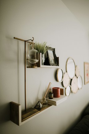 A minimalist wall shelf decorated with various items including a planter with green foliage, a framed photo, a glass terrarium, and small decorative objects like a pyramid. The shelf has a light wood finish with a gold bar supporting it. Below the shelf, a series of circular wall art pieces are arranged in an overlapping pattern. The overall setting gives a cozy and aesthetically pleasing vibe.