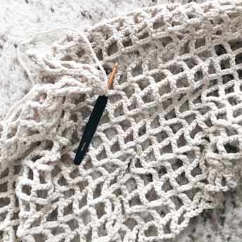 A black crochet hook is inserted in a piece of beige, open-knit fabric that is spread out on a soft, textured surface.