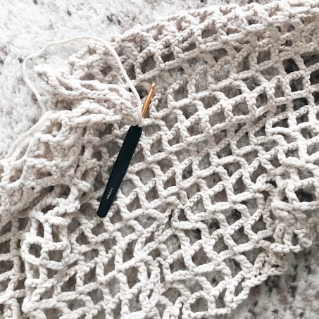 A black crochet hook is inserted in a piece of beige, open-knit fabric that is spread out on a soft, textured surface.