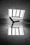 A black and white photograph of a ceiling featuring two square windows and a ceiling fan with three blades. The windows have metal frames and are open, allowing natural light to pass through. The surface of the ceiling exhibits a rough texture, adding to the industrial feel of the image.