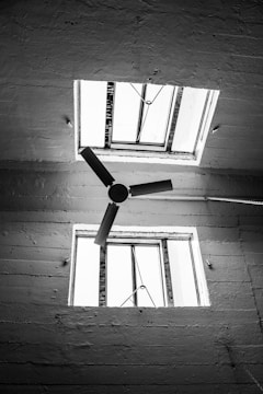 A black and white photograph of a ceiling featuring two square windows and a ceiling fan with three blades. The windows have metal frames and are open, allowing natural light to pass through. The surface of the ceiling exhibits a rough texture, adding to the industrial feel of the image.