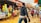 A bustling outdoor market scene with various people walking and shopping. Numerous colorful packages of goods are displayed on wooden tables, with shoppers interacting and browsing. The ground is muddy and reflects activity typical of a vibrant market environment.