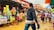 A bustling outdoor market scene with various people walking and shopping. Numerous colorful packages of goods are displayed on wooden tables, with shoppers interacting and browsing. The ground is muddy and reflects activity typical of a vibrant market environment.