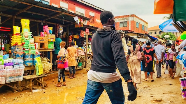 A bustling outdoor market scene with various people walking and shopping. Numerous colorful packages of goods are displayed on wooden tables, with shoppers interacting and browsing. The ground is muddy and reflects activity typical of a vibrant market environment.