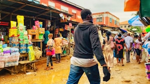 A bustling outdoor market scene with various people walking and shopping. Numerous colorful packages of goods are displayed on wooden tables, with shoppers interacting and browsing. The ground is muddy and reflects activity typical of a vibrant market environment.