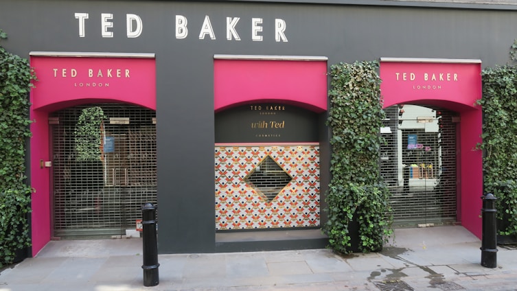 A storefront with three arched entrances featuring a vibrant, pink color. Each arch is framed by lush green ivy, and the name 'TED BAKER' is prominently displayed above. The middle arch has a patterned design with a diamond shape in the center, while the others are covered with a metal gate.