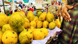 A vibrant market scene with a close-up view of a large pile of yellow and green mangoes displayed on a woven basket covered with paper. Several people in colorful clothing are visible in the background, with one person prominently holding a hand on their stomach while engaging with others.