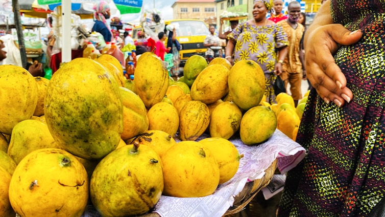 A vibrant market scene with a close-up view of a large pile of yellow and green mangoes displayed on a woven basket covered with paper. Several people in colorful clothing are visible in the background, with one person prominently holding a hand on their stomach while engaging with others.