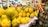 A vibrant market scene with a close-up view of a large pile of yellow and green mangoes displayed on a woven basket covered with paper. Several people in colorful clothing are visible in the background, with one person prominently holding a hand on their stomach while engaging with others.