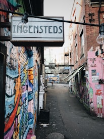A narrow alleyway is filled with vibrant street art on the brick walls, featuring various colorful designs and characters. A sign overhead reads 'Ingensteds'. The area is mostly deserted except for a lone person walking in the distance. Urban elements such as lamp posts, stairs, and other signs are visible, giving the space a gritty, artistic vibe.